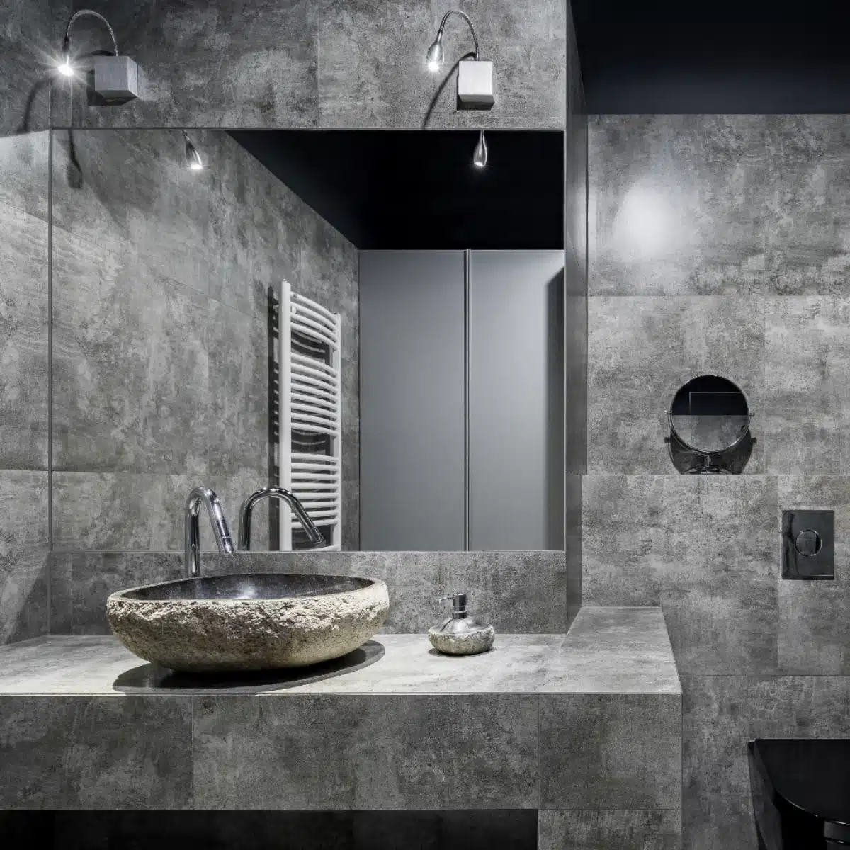 A modern bathroom with gray stone tiles, a large round stone sink, chrome faucet, wall-mounted mirror, minimalist soap dish, and a white heated towel rail in the background.