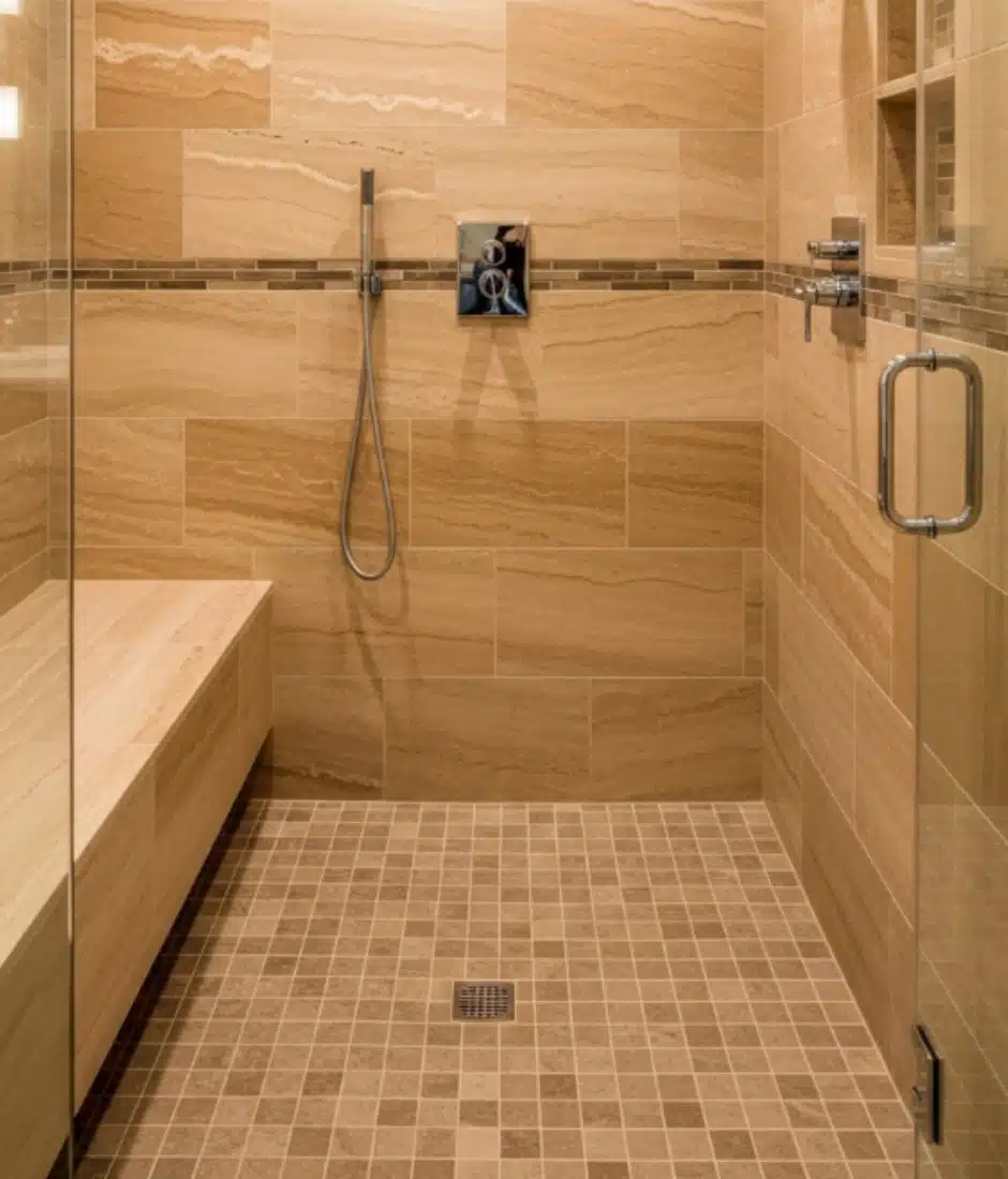 A modern walk-in shower with tan stone tile walls and floor, a built-in bench on the left, a handheld showerhead, a glass door, and a horizontal decorative tile accent on the wall.
