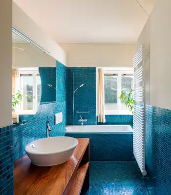 Modern bathroom with blue mosaic tiles, a white vessel sink on a wooden countertop, a bathtub with shower, large window with curtains, white radiator, and green plants visible by the window.