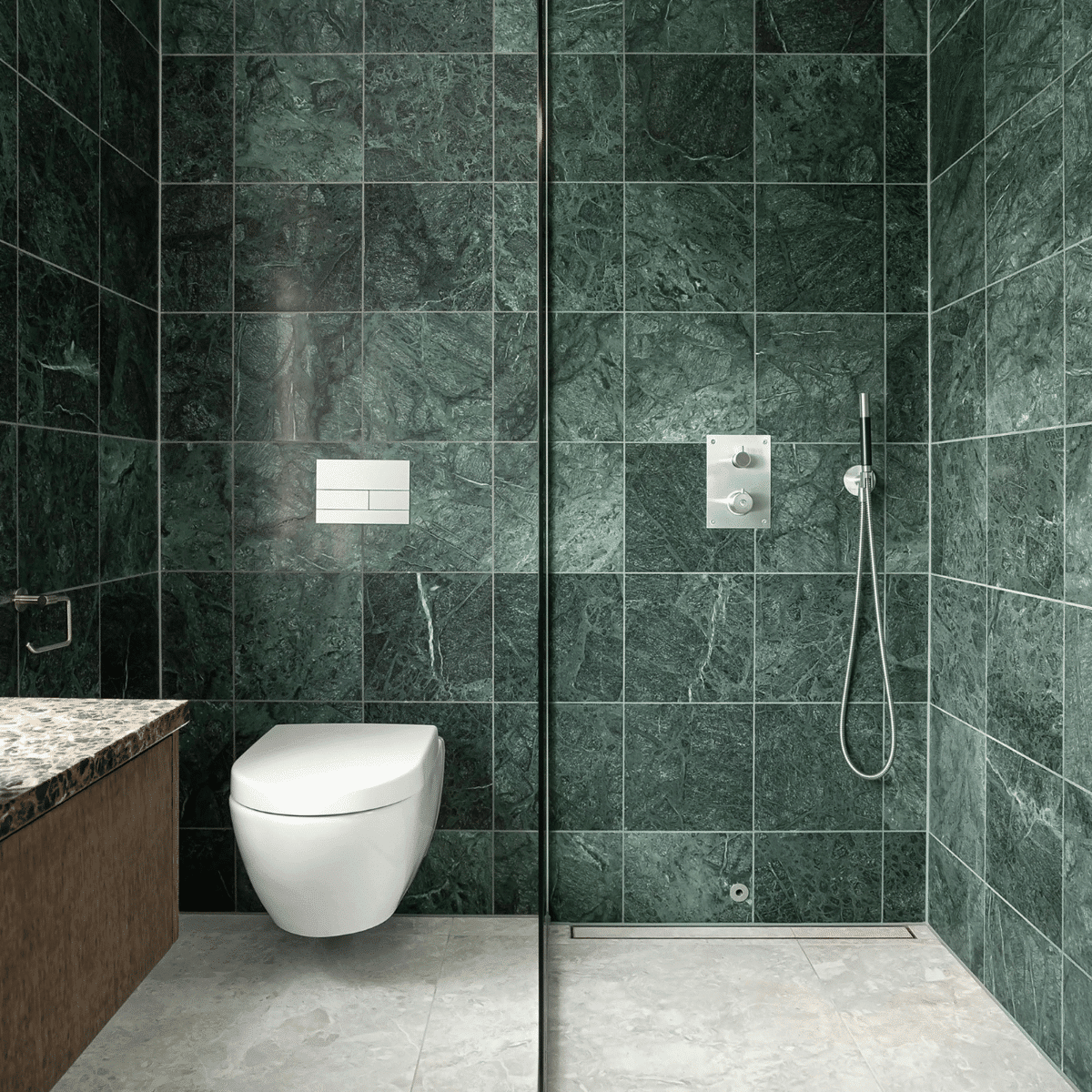 Modern bathroom with dark green marble wall tiles, a floating white toilet, a walk-in shower with a handheld showerhead, and a marble countertop vanity with wood paneling. Floor is light gray stone.