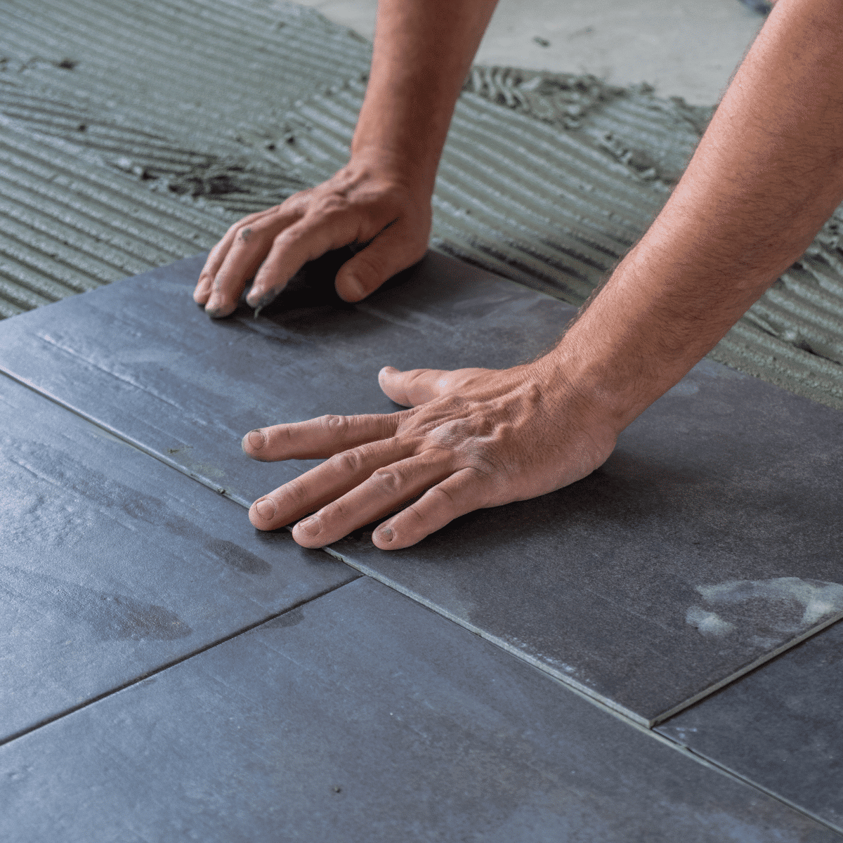 Close-up of a person’s hands installing dark gray ceramic tiles on a floor, pressing the tiles into adhesive with ridged patterns visible beneath.