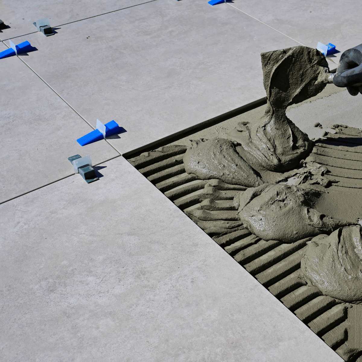 A hand spreads mortar on the floor with a trowel, preparing to lay a large tile. Other tiles are already placed, separated by blue and white spacers for alignment.