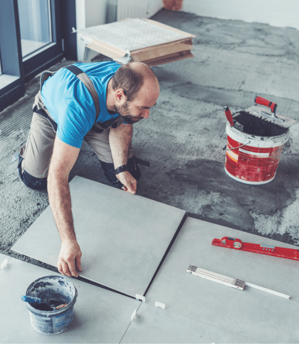 A man in work clothes kneels on the floor, carefully placing a large gray tile. Nearby are a bucket of adhesive, a red spirit level, and other tiling tools. The room appears to be under renovation.