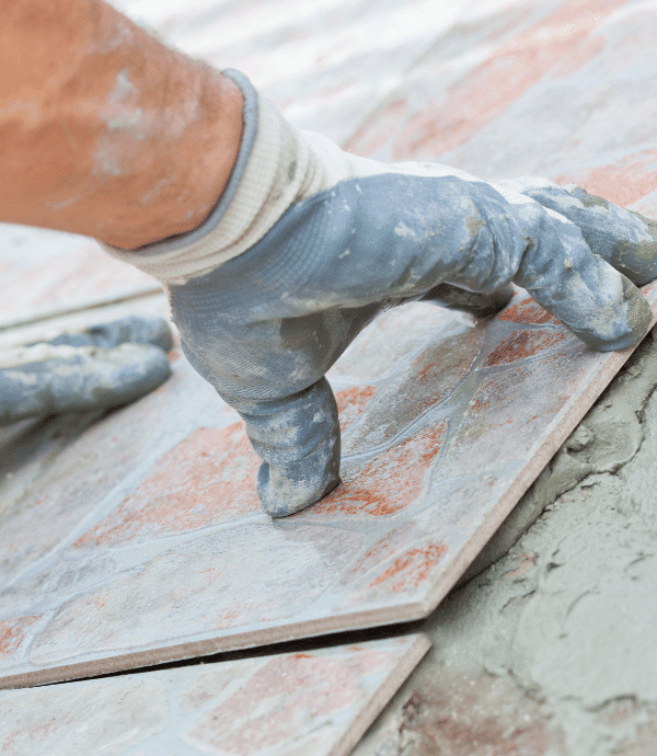 A gloved hand is placing a ceramic tile onto wet mortar during a floor tiling installation.