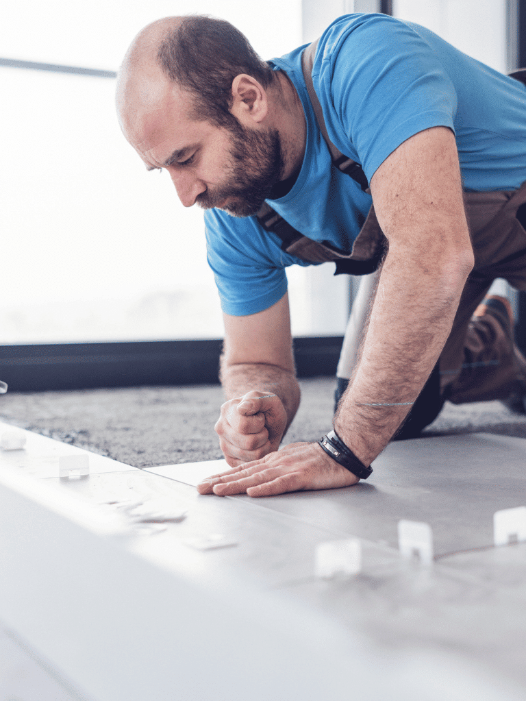 A man in work overalls and a blue shirt kneels on the floor, carefully installing large tiles and using spacers to align them, with sunlight coming through a large window in the background.