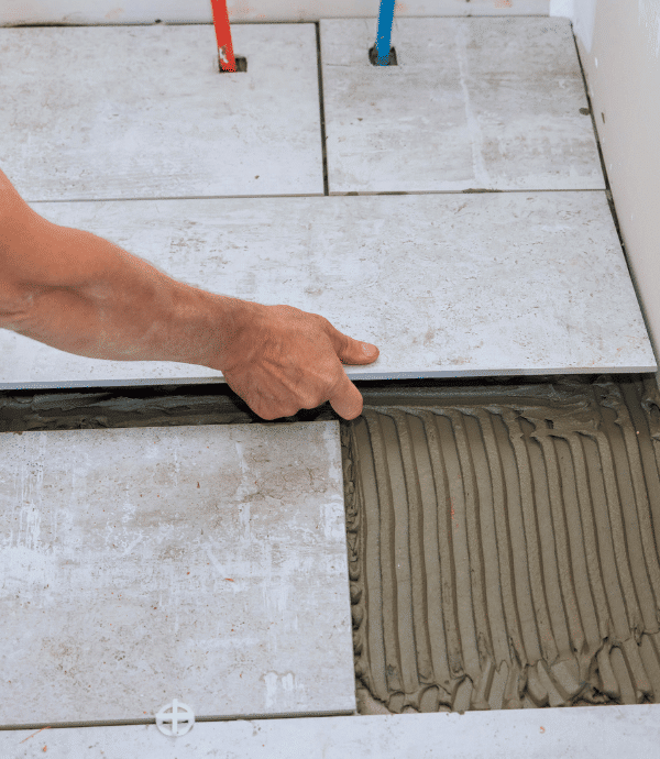 A person’s hand is placing a ceramic tile onto a floor with visible adhesive underneath. Several tiles are already laid, and two pipes protrude from the wall in the background.
