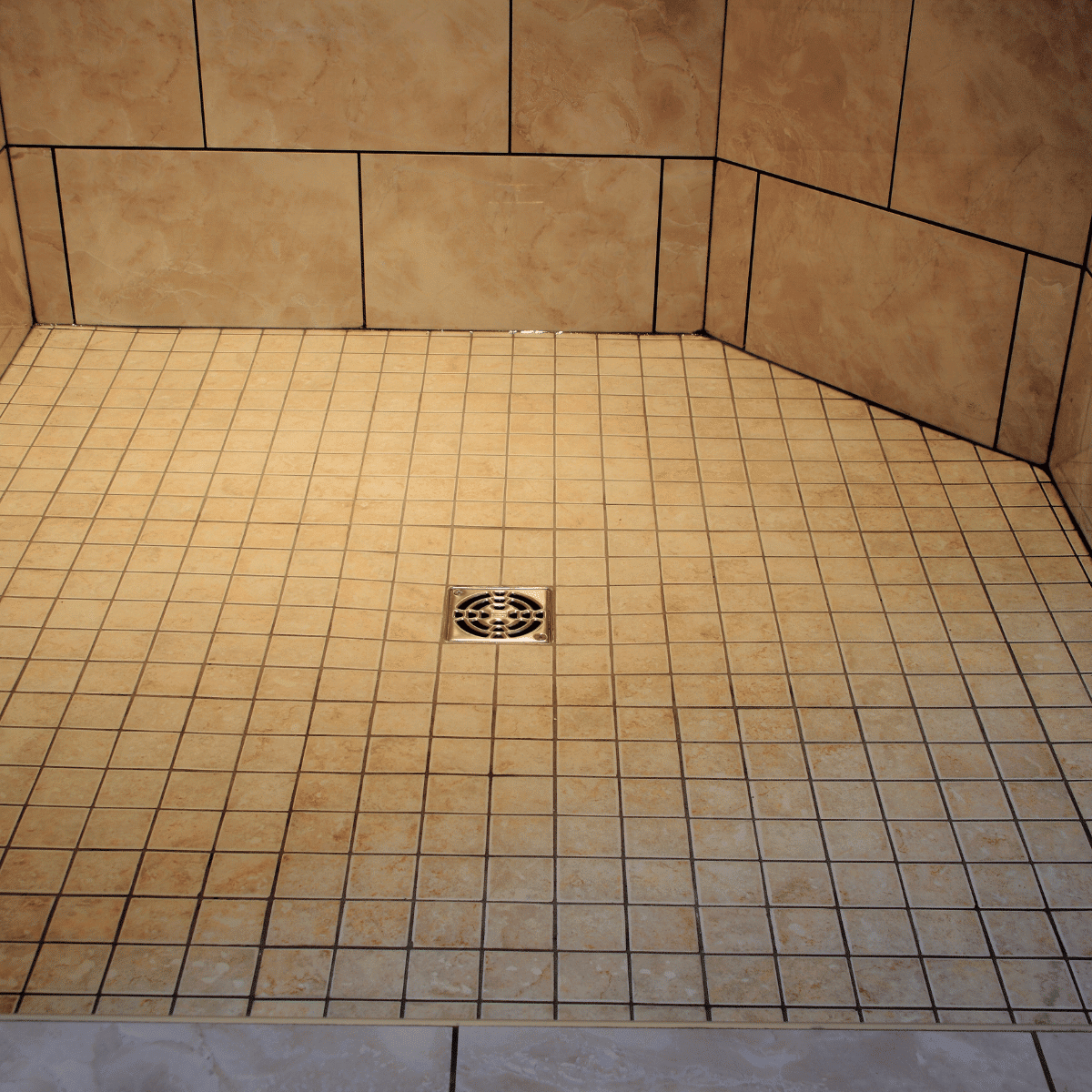 A tiled shower floor with beige square tiles, grout lines, and a silver drain in the center, surrounded by matching tiled walls.