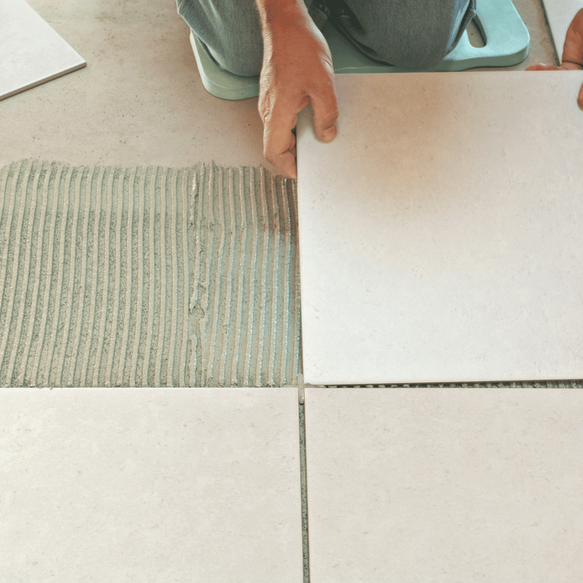 A person installs large, light-colored ceramic tiles on a floor, pressing one tile into adhesive with their hands while kneeling.