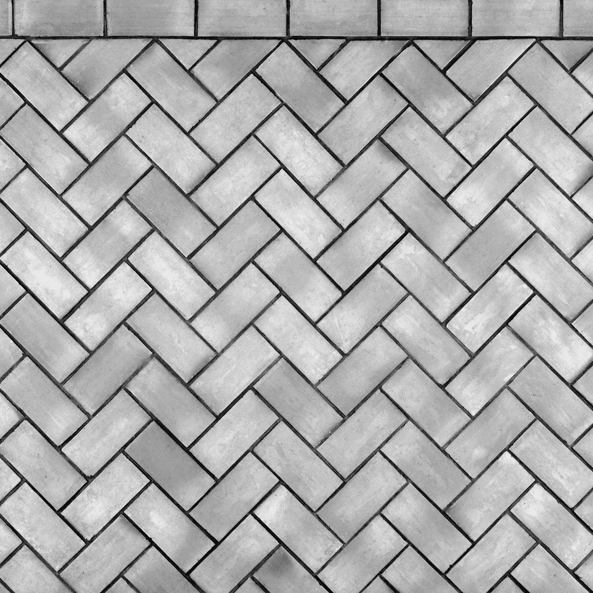 Gray rectangular tiles arranged in a herringbone pattern, with a straight row of tiles forming a border at the top of the image. The tiles have slight variations in shading.