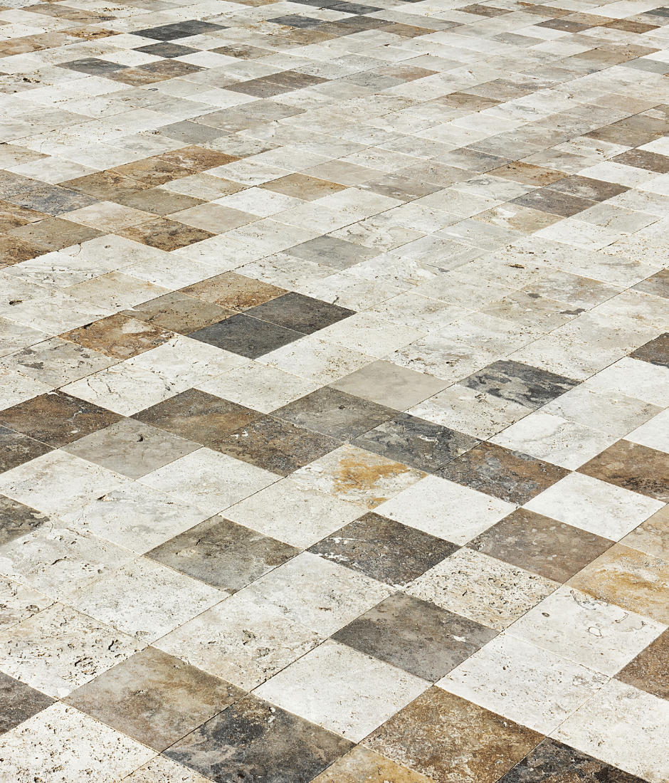 A tiled floor made of square stone tiles in various shades of beige, brown, and gray, creating a checkerboard pattern with a natural, textured appearance.
