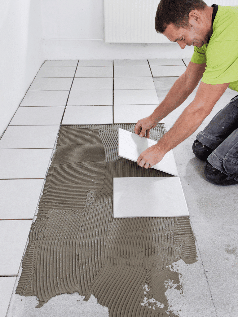 A person in a green shirt is kneeling and placing floor tiles onto a layer of adhesive, carefully aligning each tile to match the existing tiled area.