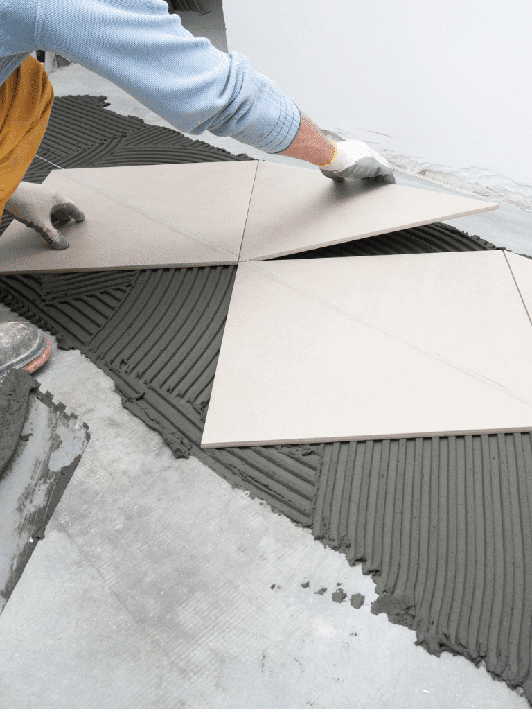 A person is laying large square tiles onto a floor covered with wet tile adhesive, carefully aligning the tiles with gloved hands.